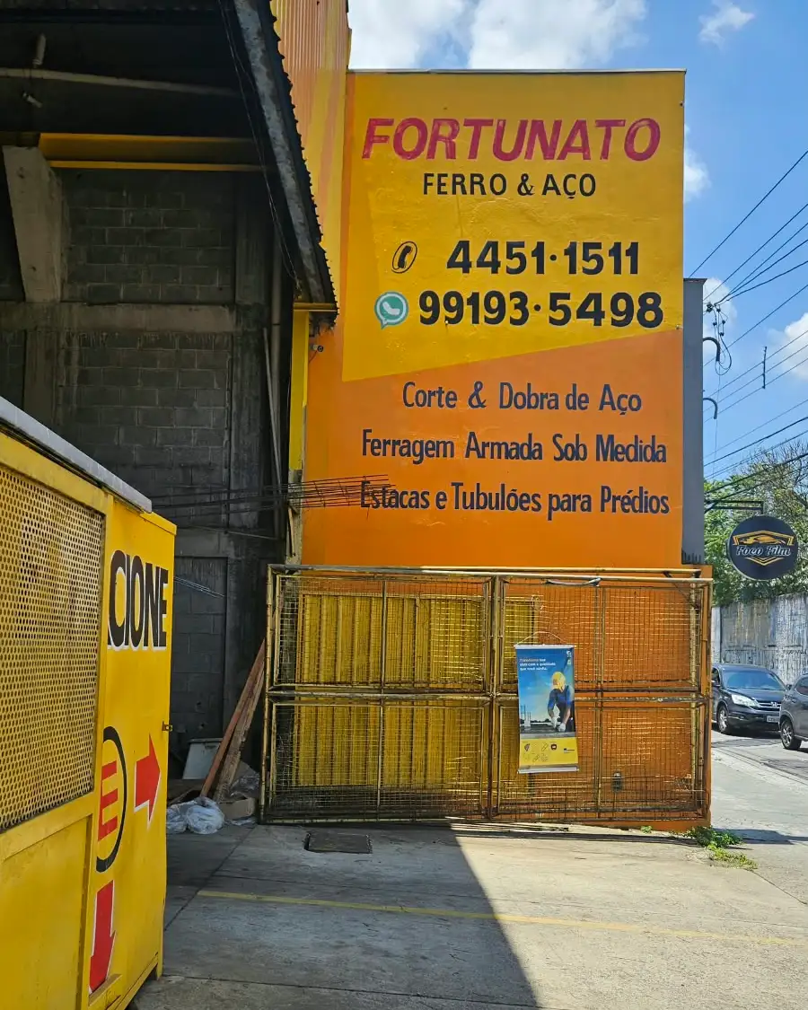 Muro amarelo da loja com o nome Fortunato Ferro e Aço, os telefones e os serviços: corte e dobra de aço, ferragem armada sob medida, estacas e tubulões para prédios
